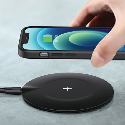 Dłoń trzyma smartfon Apple iPhone nad czarną ładowarką indukcyjną Usams US-CD149, na której widnieje biały symbol plusa, podczas procesu bezprzewodowego
