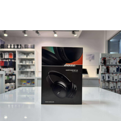 Czarne słuchawki bezprzewodowe Bose QuietComfort SC na opakowaniu z logo marki, widok na profil urządzenia z przyciskami sterującymi na tle sklepu.